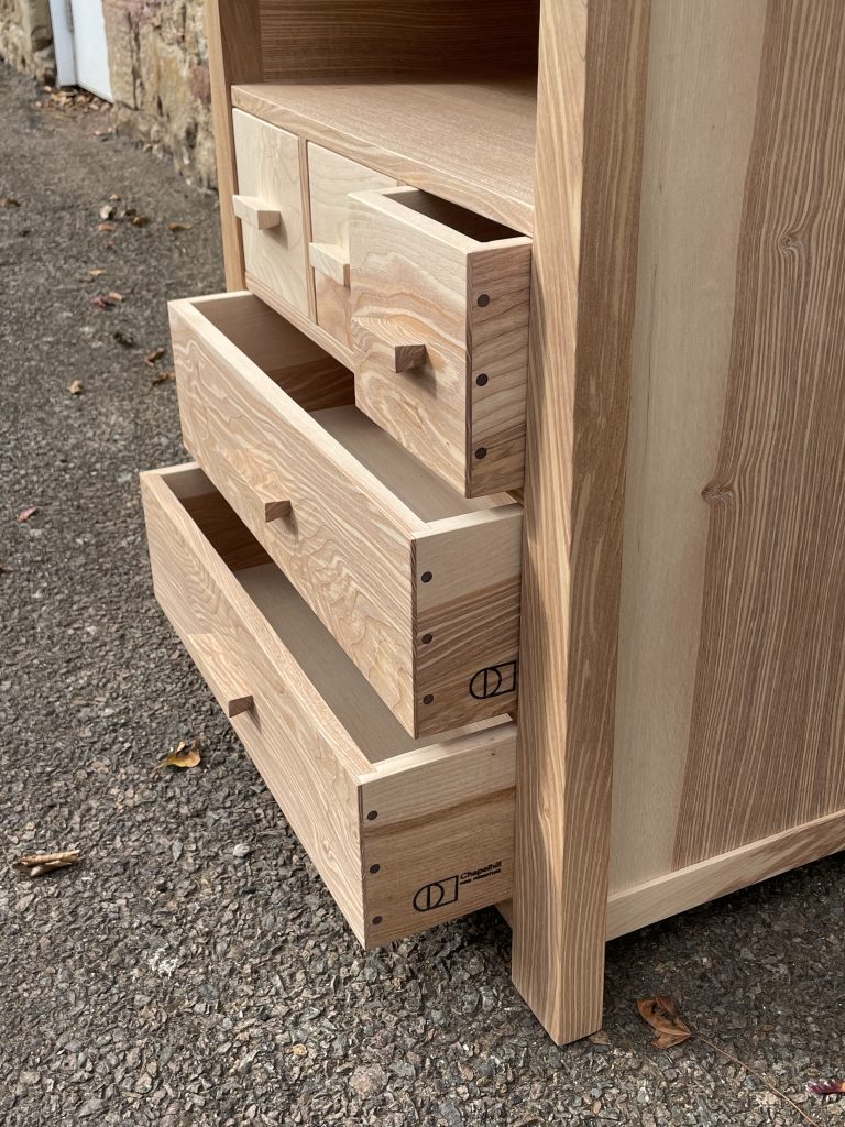 "Chaffinch" bedside cabinet made by Iain Stirling of Chapelhill Fine Furniture. Close-up side view of drawers showing walnut contrasting pegs and Chapelhill branding.
