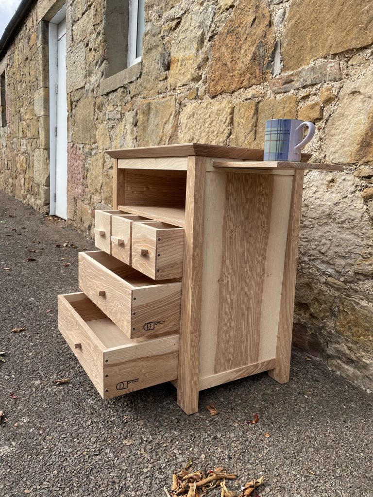 "Chaffinch" bedside cabinet made by Iain Stirling of Chapelhill Fine Furniture. View from right with mug on tray and drawers open.