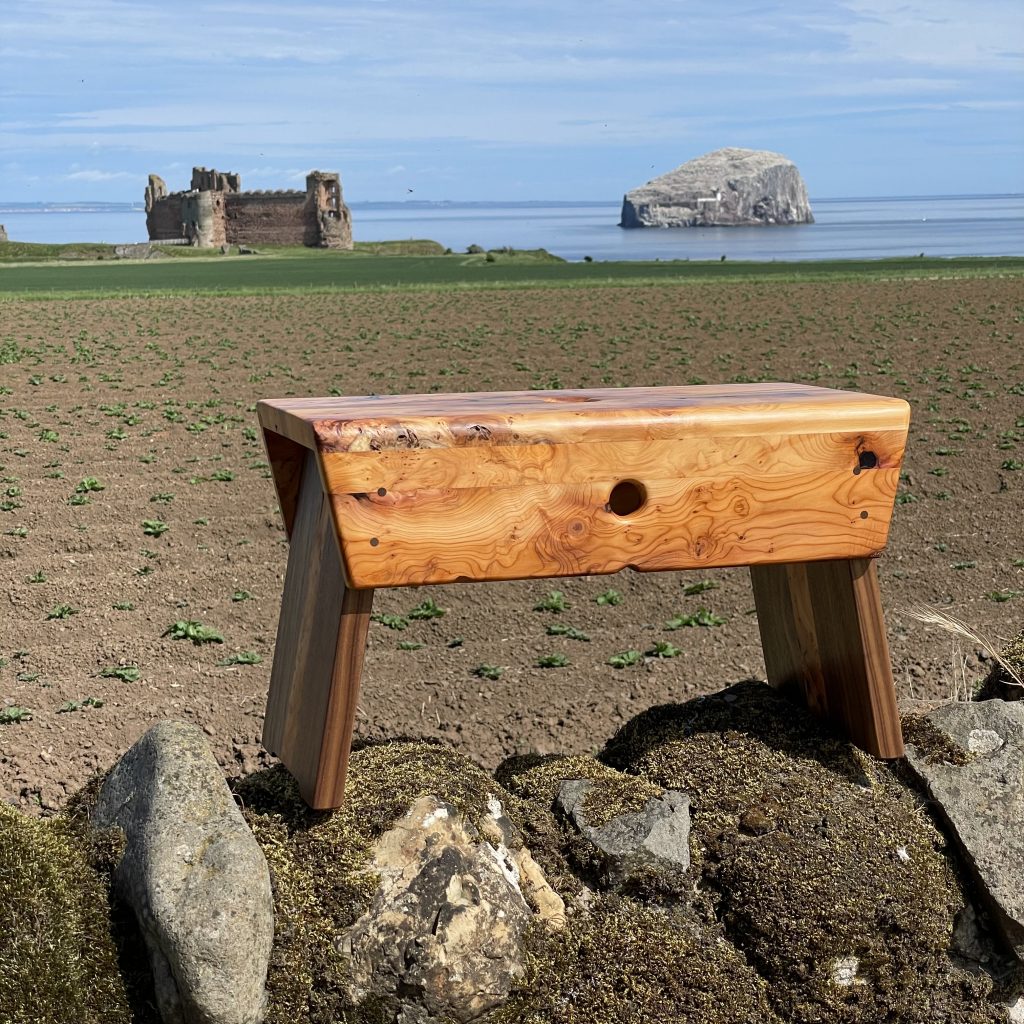Yew and walnut Scottish creepie stool made by Chapelhill Fine Furniture in Scotland, with Tantallon Castle and Bass Rock in background.