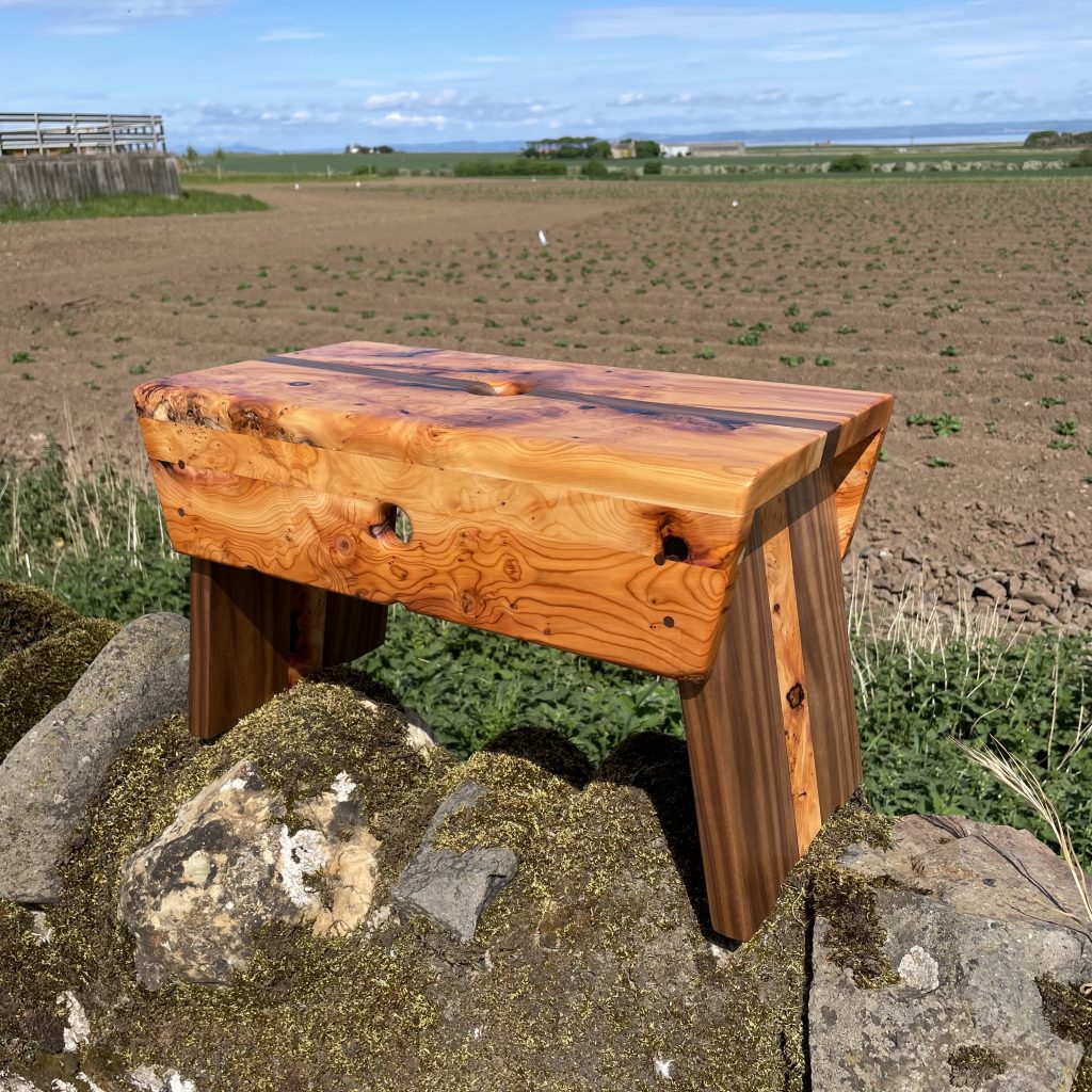 Yew and walnut Scottish creepie stool made by Chapelhill Fine Furniture in Scotland, with Tantallon Castle and Bass Rock in background.