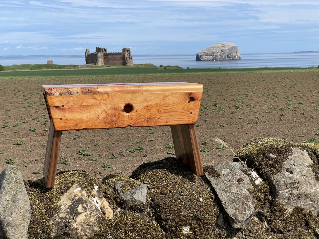 Yew and walnut Scottish creepie stool made by Chapelhill Fine Furniture in Scotland, with Tantallon Castle and Bass Rock in background.